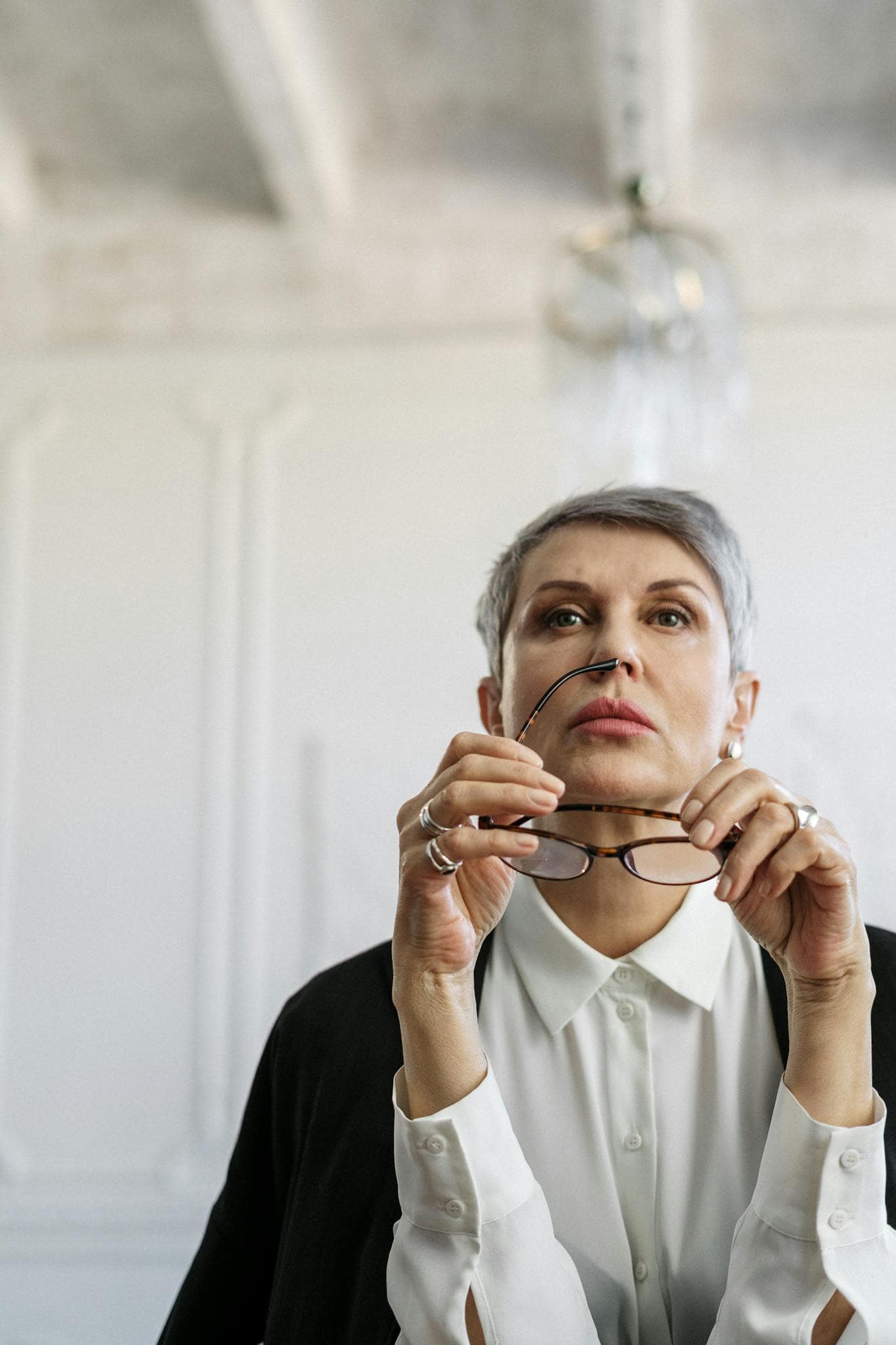 Thoughtful senior woman holding glasses, reflecting in a bright, modern interior setting.